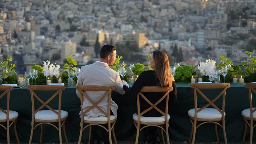 A couple sits at an outdoor table topped with floral arrangements while looking out at the city below