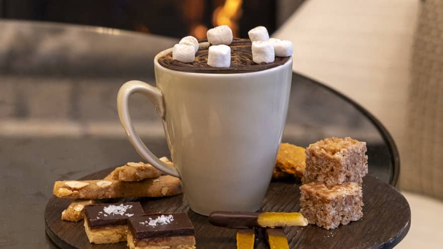 White mug of hot chocolate with mashmallows on a dark, round platter filled with small desserts