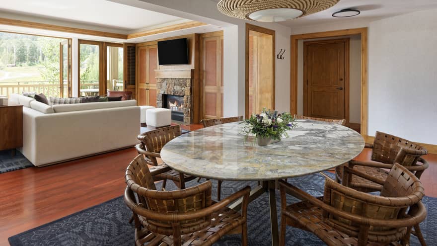 Luxury suite with a marble dining table, woven chairs, sectional sofa, stone fireplace, TV and large windows opening to a scenic balcony view