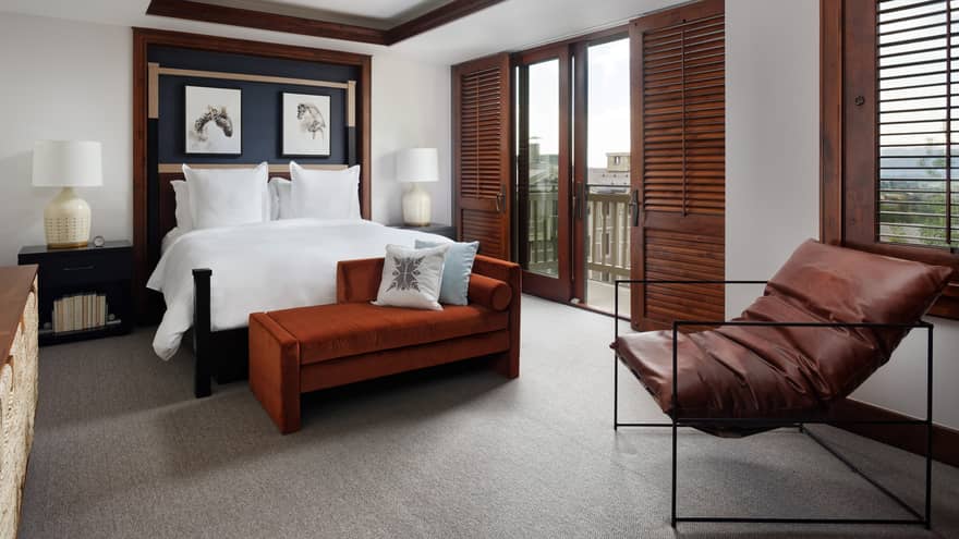Luxury resort bedroom with a king bed, nightstands with lamps, burnt orange chaise, leather lounge chair and dark wooden doors opening to a balcony view