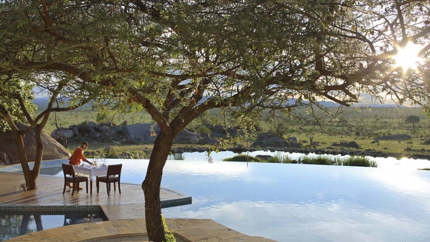 Hotel staff sets small patio table on wood platform beside tree, outdoor swimming pool
