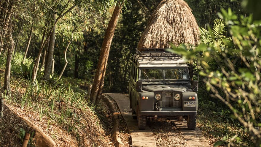 Jeep driving down path in forest past thatched-roof hut