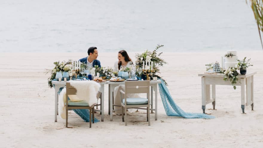 Smiling couple at private dining on white sand beach past palm leaves