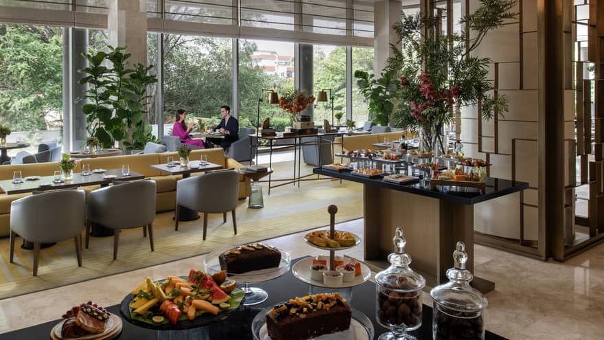 Two people sit a small table by window in room with metal d�cor and various types of food on table display