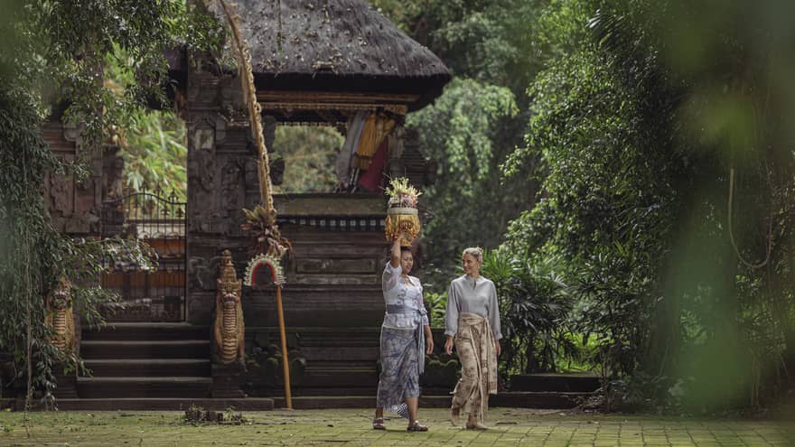 Two women wearing batik skirts walk by a temple in a lush tropical area as one of them carries a golden tray on her head.