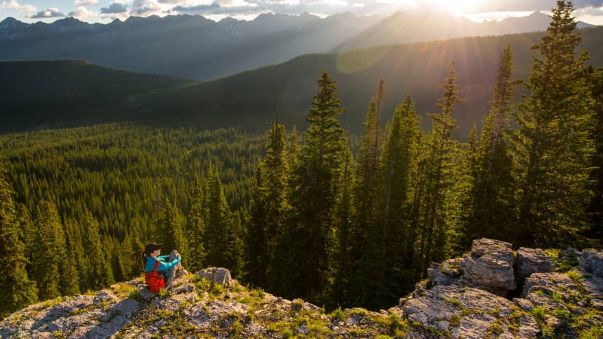 Person sits at edge of cliff overlooking trees, mountains as sun starts to set