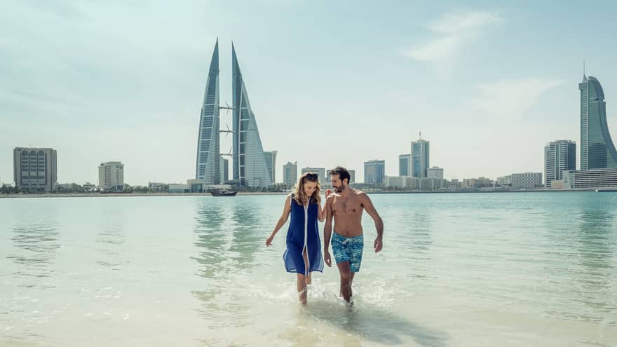 A man and woman run arm in arm through the shallow ocean water with Bahrain Bay skyscrapers in the background