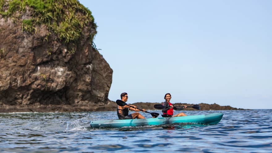 Two people paddle in a bright blue kayak through the ocean, passing by a large rock outcropping
