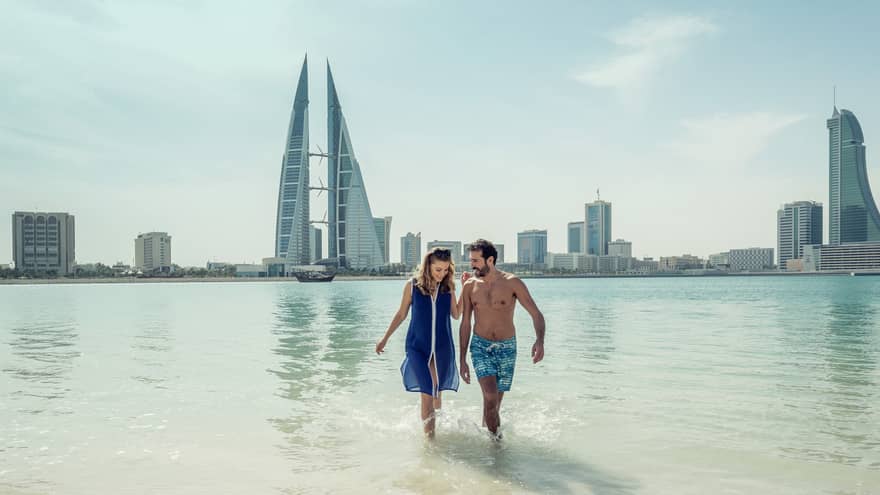 A man and woman run arm in arm through the shallow ocean water with Bahrain Bay skyscrapers in the background