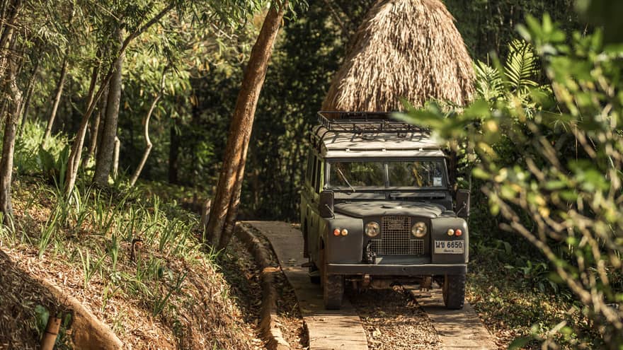 Jeep driving down path in forest past thatched-roof hut