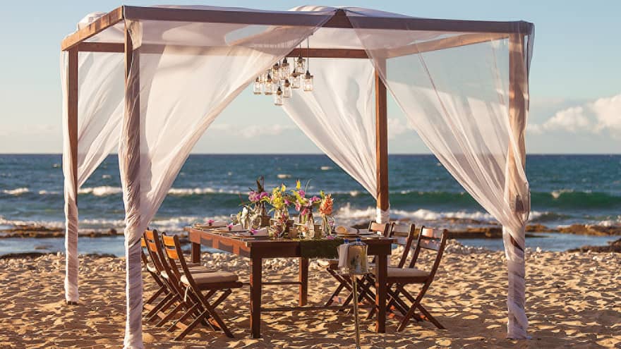 Wood dining table with fresh flowers under sheer white canopy, lanterns on beach