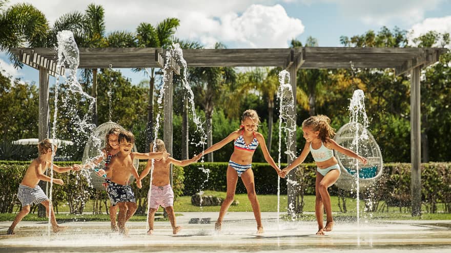 Group of young children in swimsuits runs across outdoor splash pad with fountains