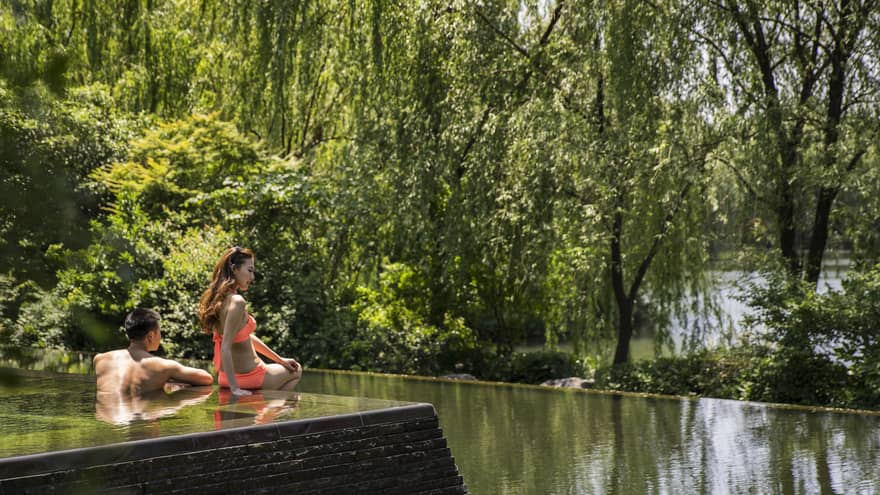Woman in orange bikini sits on edge of swimming pool in front of man