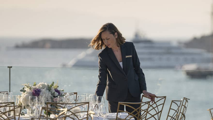 Woman arranges round table with glasses, flowers, white and gold chairs overlooking ocean and boat