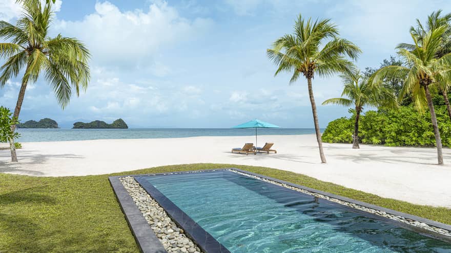 Rectangular in-ground pool jutting out to white-sand beach dotted with palm trees with two lounge chairs and umbrella along blue waters