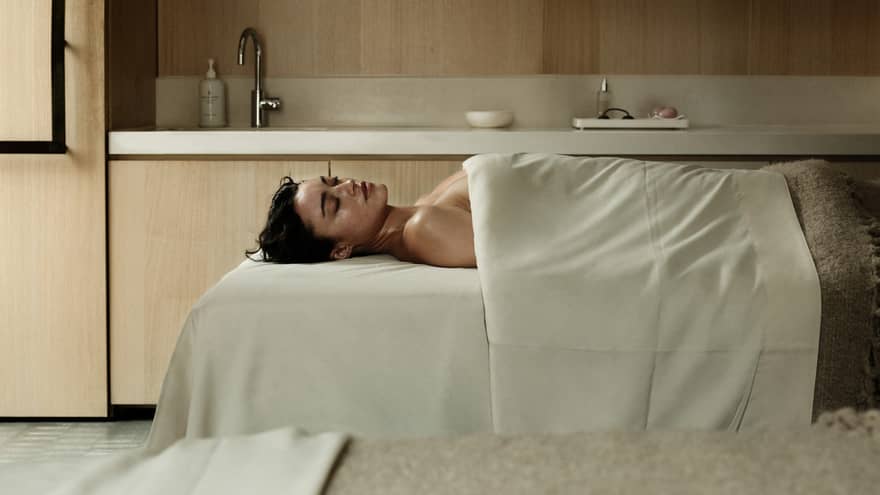 A person lies on supine on a spa treatment table in a serene, spacious treatment room and is draped with with a sheet and blanket.