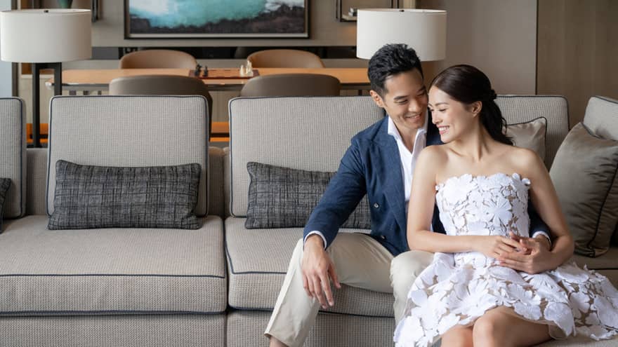Smiling couple seated on a light gray sofa in a modern living room with decorative shelves and a large ocean artwork in the background.