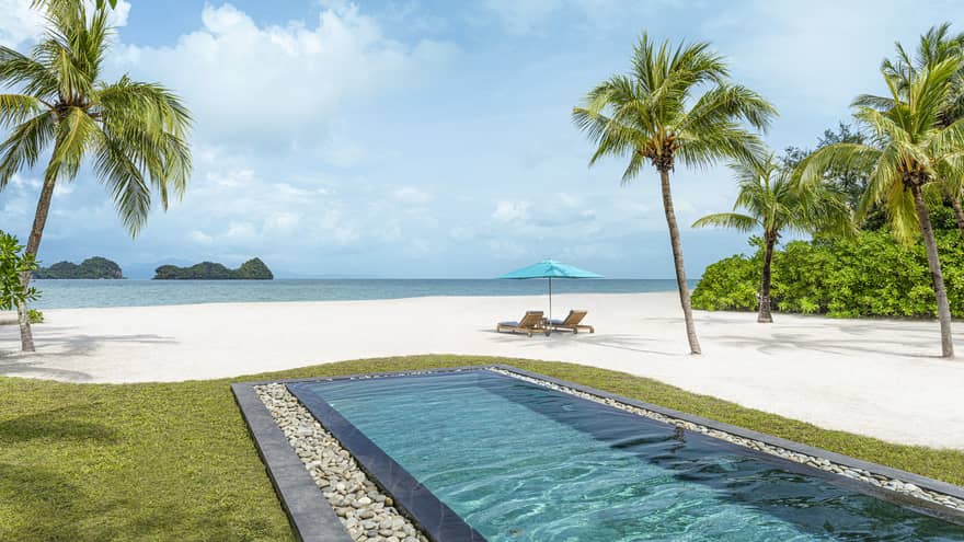 Rectangular in-ground pool jutting out to white-sand beach dotted with palm trees with two lounge chairs and umbrella along blue waters