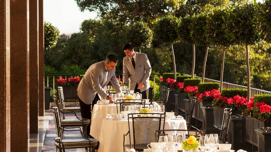 Two servers in grey jackets with black ties set outdoor dining tables with white tablecloths, dishes, red flowers along side