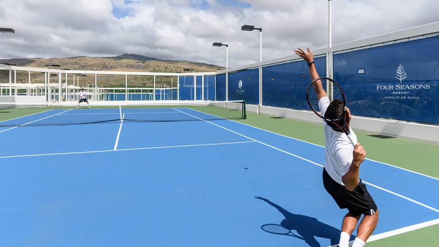 View from the court's back corner of a player about serve the ball on an outdoor tennis court, mountains in the distance.
