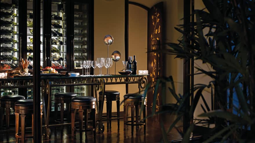 Wine bottle, glasses, cheese platter on table in front of wine cellar in dark restaurant