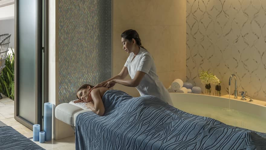 A woman under a blue sheet gets a massage from a masseuse surrounded by candles and natural light. 
