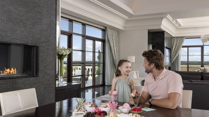 Man and young girl sit at dining table in hotel suite, next to fireplace