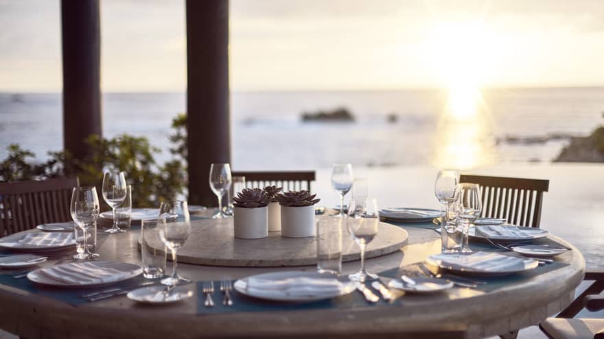 Round outdoor dining table with wine glasses, ocean and sunset in background
