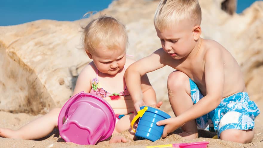 ,Young blonde boy and blonde toddler girl in swimsuits add sand to buckets on the beach