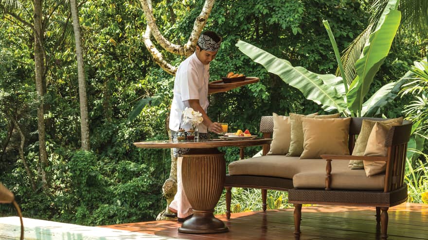 Server sets orange juice glass on in-villa dining table on patio under umbrella, villa door visible