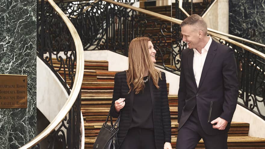 Man and woman dressed in black suits walk down wood and marble staircase
