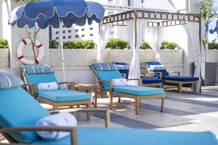 Wooden lounge chairs with team cushions, a pergola with white curtains drawn back and a blue scallop-edge umbrella on a pool deck