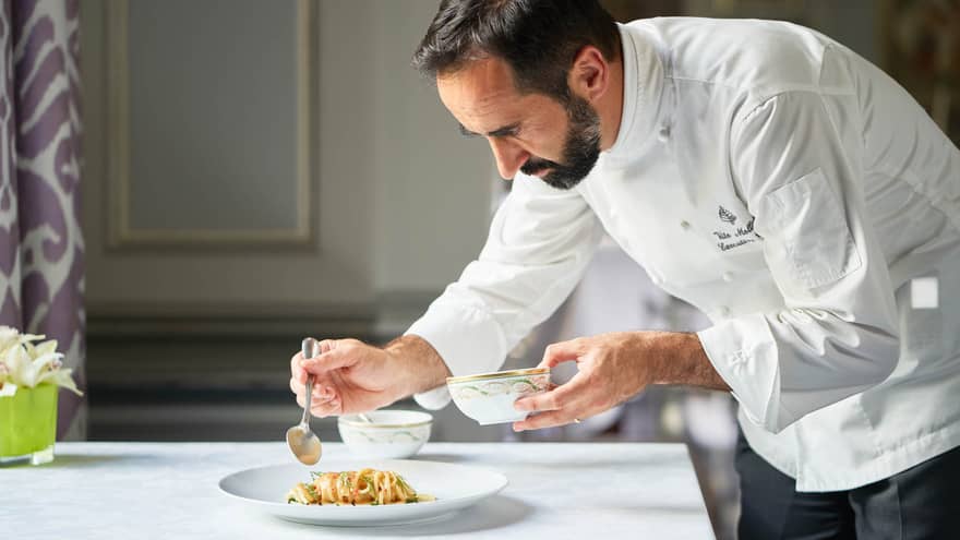 Chef Vito Mollica wearing a white chef's coat, leaning over and drizzling sauce on a plated dish