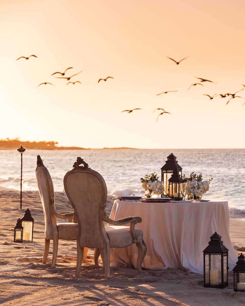 Romantic beachside dining setup with vintage chairs, a table decorated with flowers and lanterns and birds flying overhead