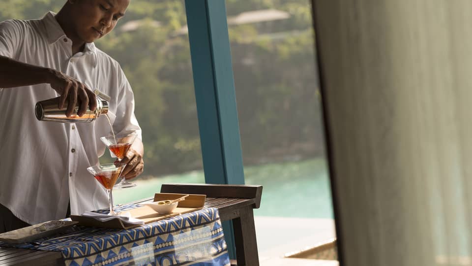 Man in white uniform with silver martini shaker pours pink cocktails into glasses on wood tray