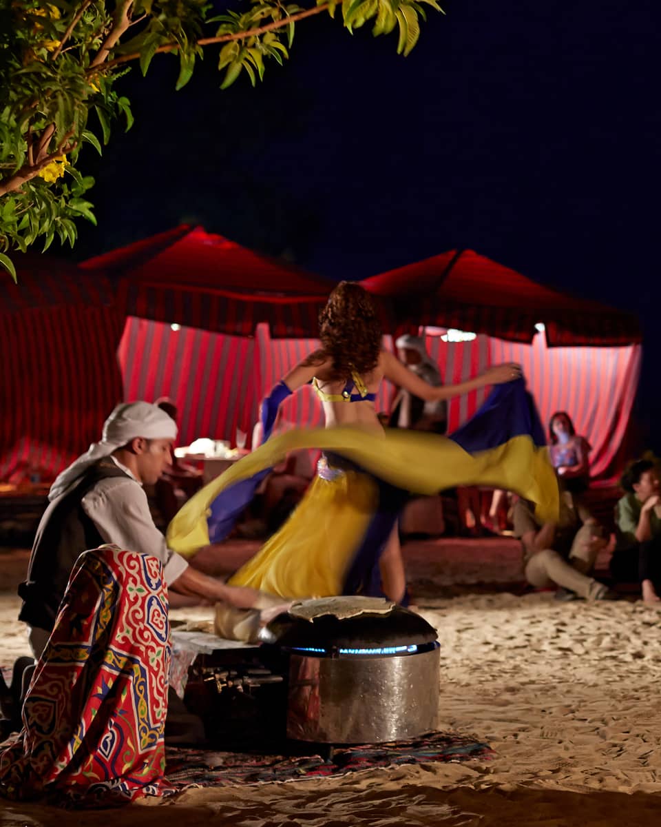 At night, a dancer performs outdoors for guests, swirling their skirt to music played by three smiling, seated musicians.