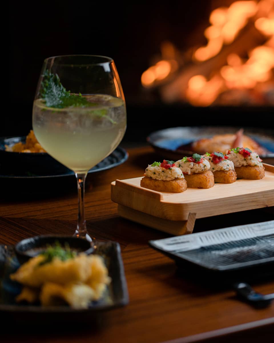 A cocktail and appetisers placed on a wooden table with a fireplace glowing in the background, creating a cosy dining atmosphere