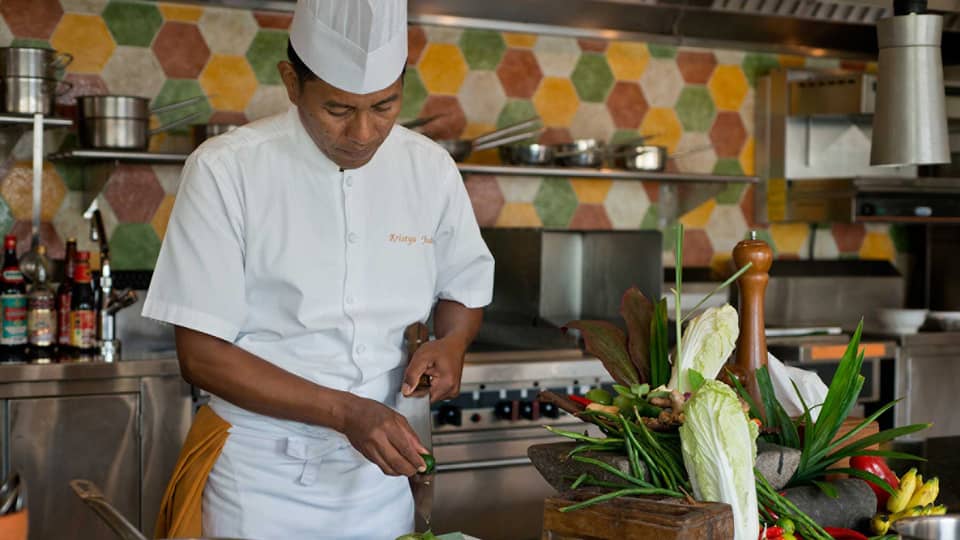 Chef in white uniform squeezes lime on large kitchen knife over dish, next to heads of fresh lettuce and whole peppers