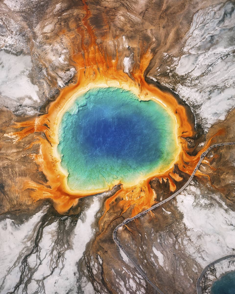 Aerial view of the Grand Prismatic Spring in Yellowstone, featuring vibrant blue, green, yellow and orange concentric rings, surrounded by rocky terrain