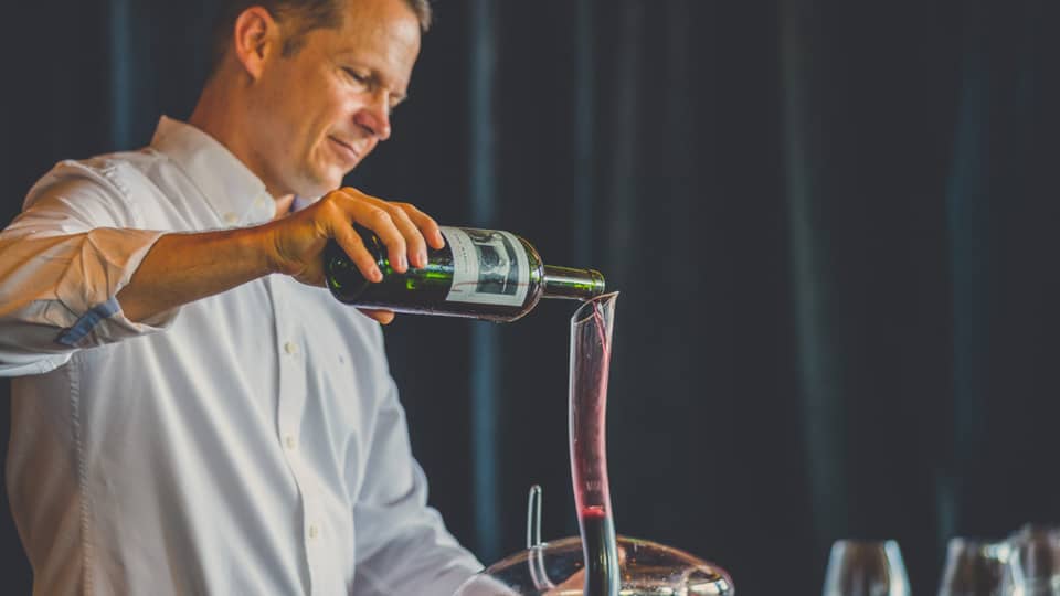 Man pours red wine into long, curved glass carafe near wine glasses