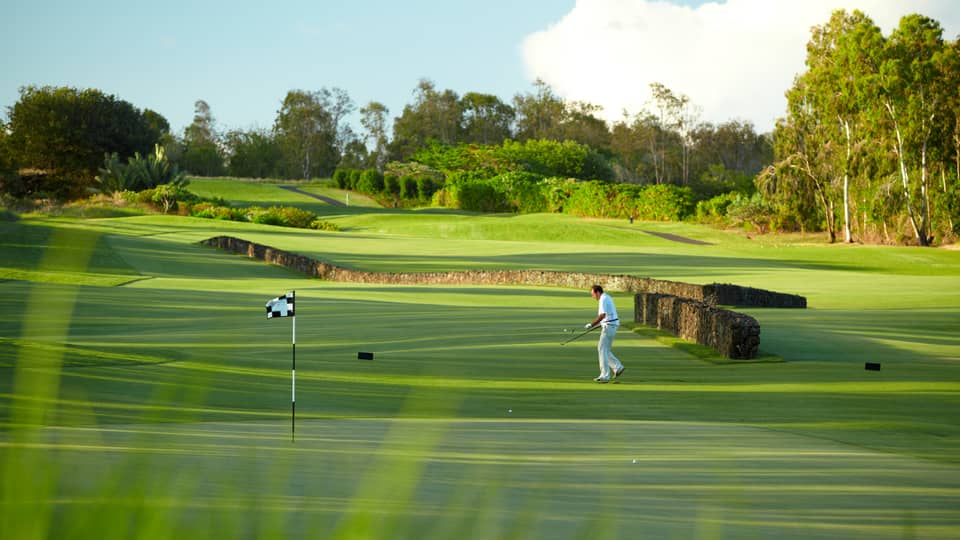 Man with golf club walks toward flag on golf course green near stone fence