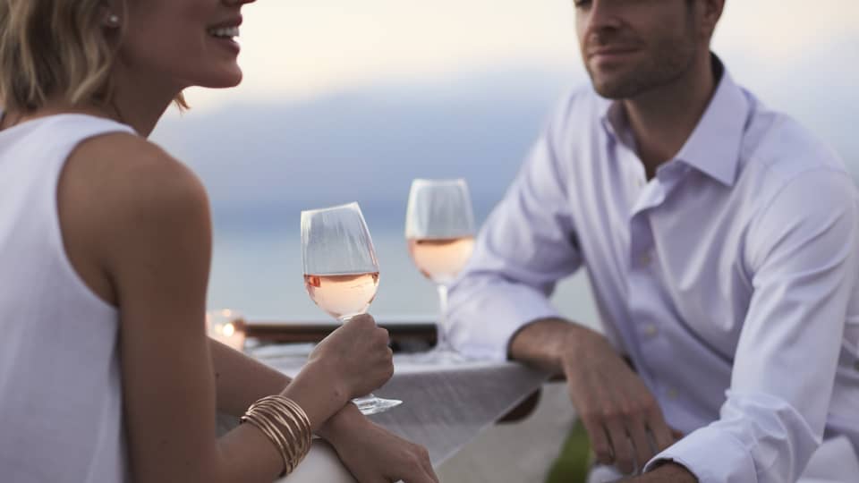 Close-up of woman and man in white, sitting and holding glasses of rose wine at dusk