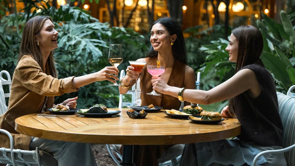 Three guests sit around a round wood table in an outdoor dining space and raise their glasses in a toast.
