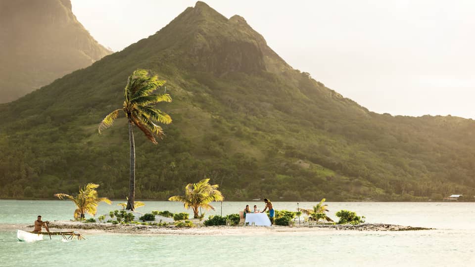 Small tropical island with palm trees, where two people are dining at a table by the water, with a mountain in the background