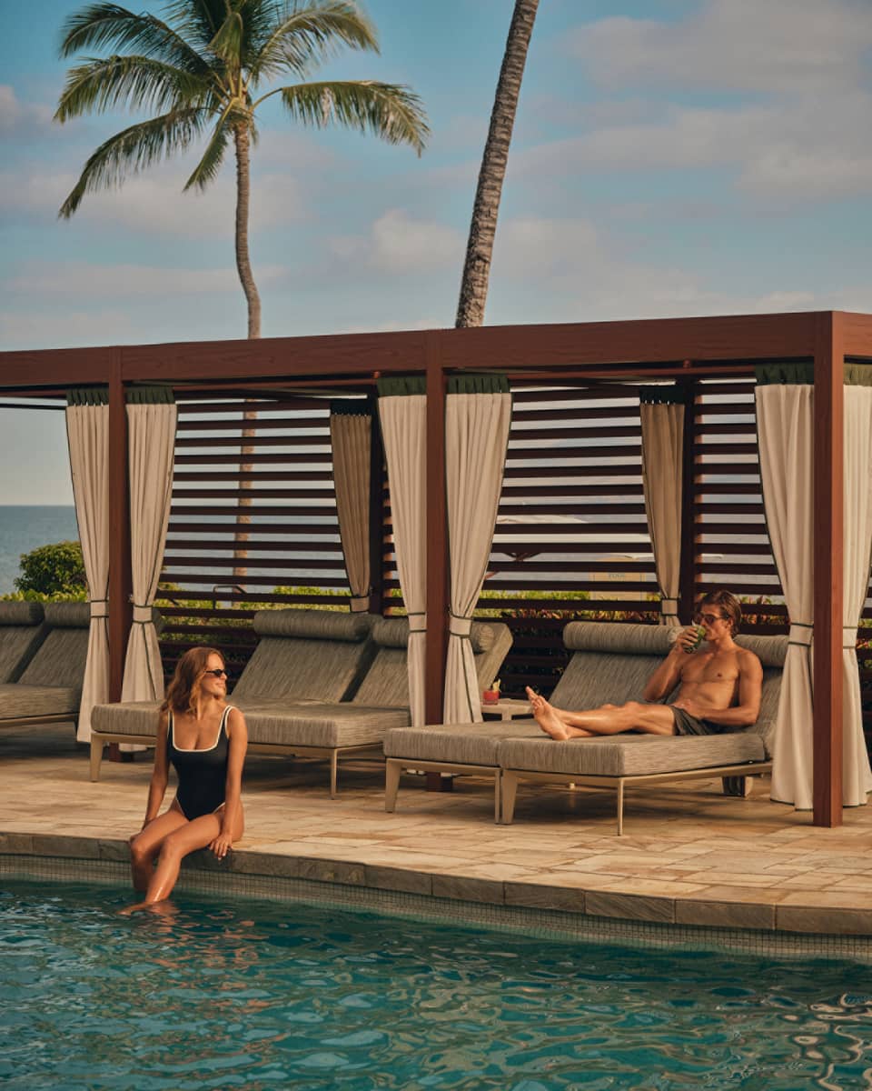 A woman sitting on the side of a pool and a man resting on a lounge chair in a cabana, with palm trees in the background