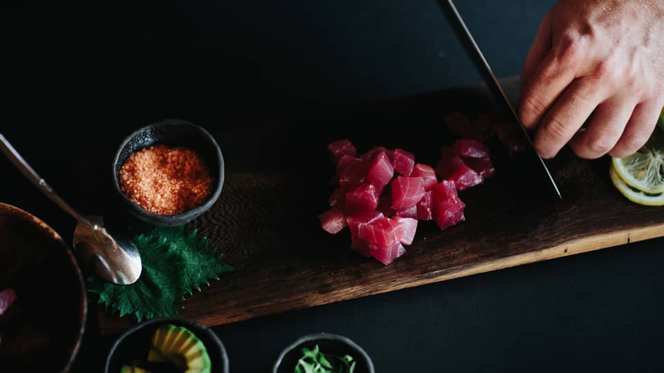 Man slicing raw tuna into cubes with knife on wood cutting board next to bowl of orange rock salt 