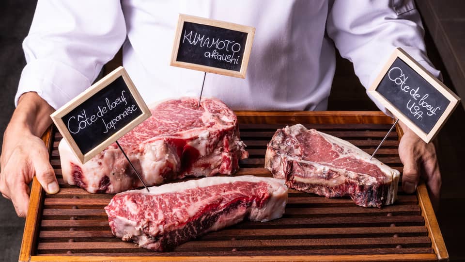 Close-up view of a wood tray with three labelled cuts of well-marbled beef, held by person in white chef's coat.