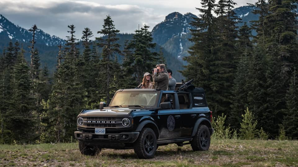 Group of friends with binoculars stand in open-top Ford Bronco surrounded by trees and mountains