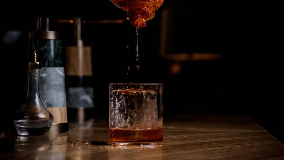 Bartender's hand pours a beverage out of a mixing glass onto a suspended croissant as it drips down into a rocks glass below