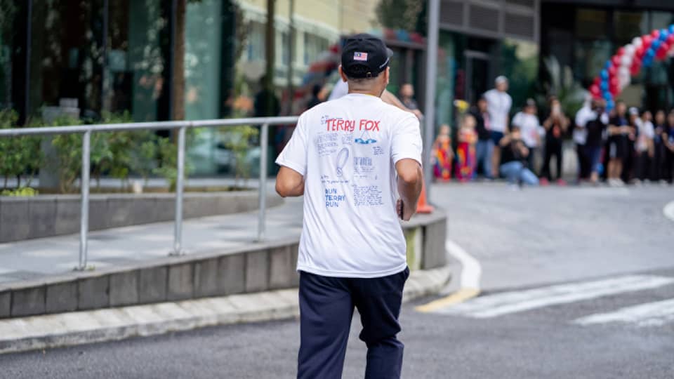 Person in white t-shirt, baseball cap and navy blue pants runs toward crowd of people under balloon arch